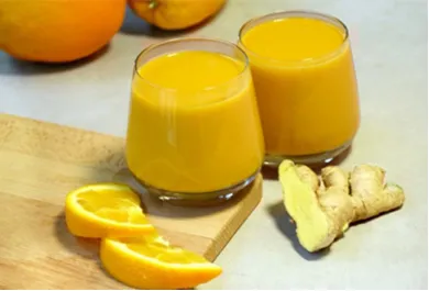 Zwei Gläser gefüllt mit orangenem Saft, daneben Orangen, Orangenscheiben und aufgeschnittener Ingwer