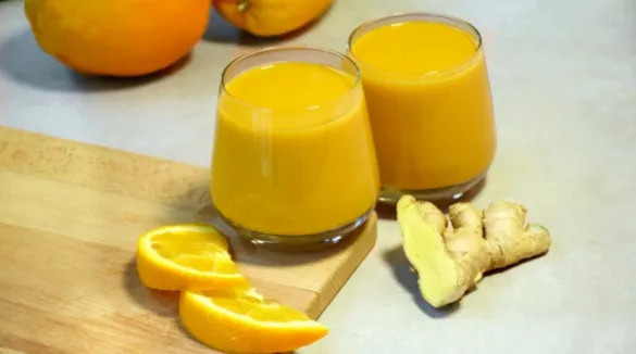 Zwei Gläser gefüllt mit orangenem Saft, daneben Orangen, Orangenscheiben und aufgeschnittener Ingwer