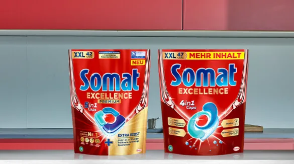 Zwei neue XXL-Packungen von Somat Excellence Geschirrspüler-Kapseln auf einer Küchenarbeitsplatte vor einem roten Hintergrund. Links ist die ‘5 in 1’ Variante mit 42 Kapseln und rechts die ‘4 in 1’ Variante mit 47 Kapseln und dem Hinweis auf mehr Inhalt zu sehen. Die Verpackung ist hauptsächlich rot mit blauen und weißen Akzenten sowie Abbildungen der Kapseln und Blasen zur Darstellung der Reinigungskraft.