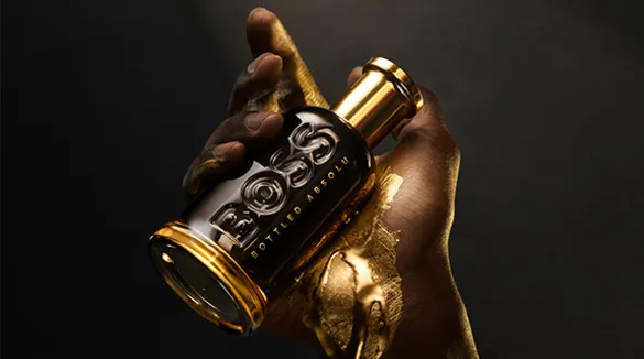 Eine mit flüssigem Gold überzogene Herrenhand hält eine Parfümflasche BOSS Bottled Absolu vor einem dunklen Hintergrund