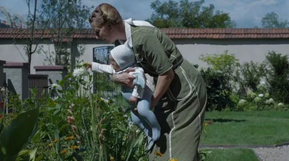 Eine Frau beugt sich mit ihrem Kind im Arm im Garten zu den Blumensträuchern vor