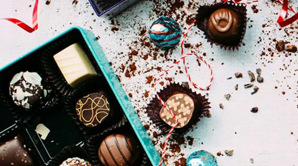 Flatlay-Pralinenpackung-türkis-rotes-Geschenkband-Schokoladenkrümel-weißer-Hintergrund