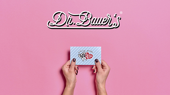 Auf rosa Hintergrund das Logo von Dr. Bauer's. Darunter Hände mit schwarz lackierten Nägeln, die eine blaue Visitenkarte mit der Aufschrift "Zahnliebe" halten